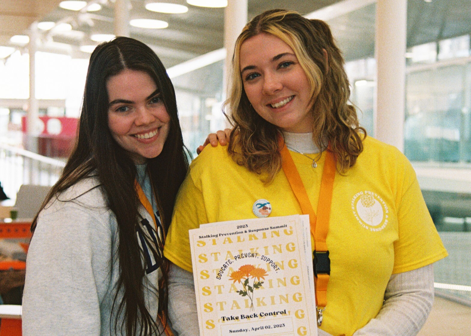 A woman with long brown hair and a gray sweatshirt and woman with wavy blonde hair wearing a yellow shirt smile at the camera.  The blonde woman is displaying a program for the Illinois Stalking Prevention and Response Summit.  