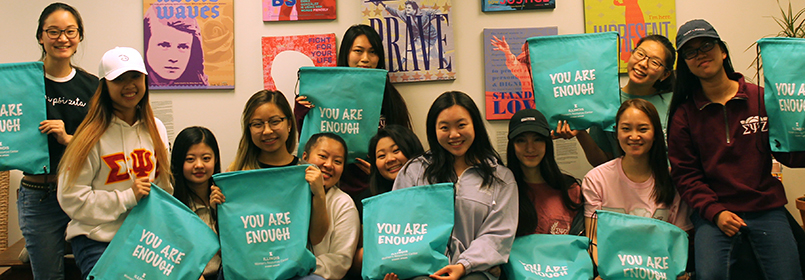 Group of 12 women holding green "You Are Enough" bags with inspirational posters hanging on back wall