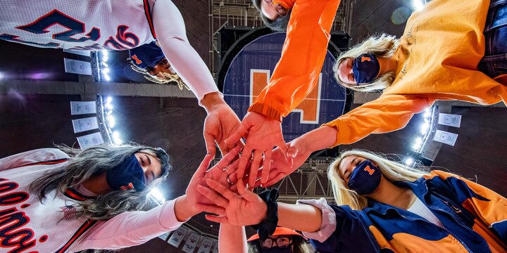 five women wearing Illinois gear put their hands together in a circle