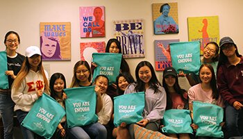 Group of 12 women holding green "You Are Enough" bags with inspirational posters hanging on back wall